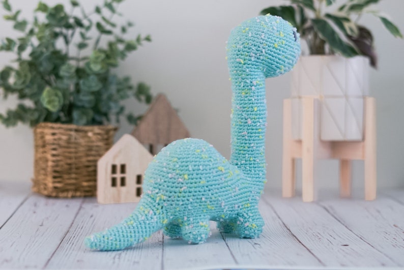 Puede incluir: Un juguete de dinosaurio de crochet azul claro con detalles blancos y amarillos. El dinosaurio est&aacute; de pie sobre una superficie de madera blanca.