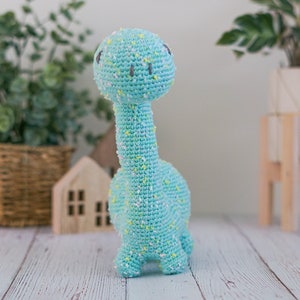 Puede incluir: Un juguete de dinosaurio de crochet azul claro con motas blancas y amarillas. El dinosaurio tiene un cuello largo y una cabeza peque&ntilde;a con ojos negros.