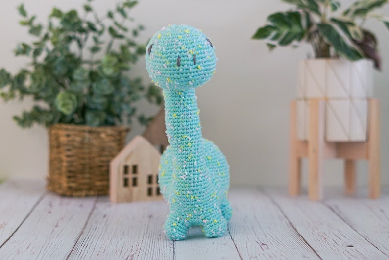 Puede incluir: Un juguete de dinosaurio de crochet azul claro con motas blancas y amarillas. El dinosaurio tiene un cuello largo y una cabeza peque&ntilde;a con ojos negros.