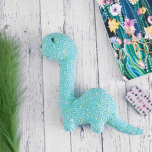 Puede incluir: Un juguete de dinosaurio de crochet azul claro con motas amarillas. El dinosaurio est&aacute; tumbado sobre una superficie de madera blanca con plantas verdes y un cuaderno con un patr&oacute;n de cactus en el fondo.