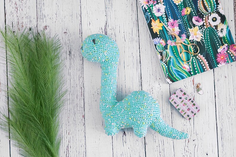 Puede incluir: Un juguete de dinosaurio de crochet azul claro con motas amarillas. El dinosaurio est&aacute; tumbado sobre una superficie de madera blanca con plantas verdes y un cuaderno con un patr&oacute;n de cactus en el fondo.