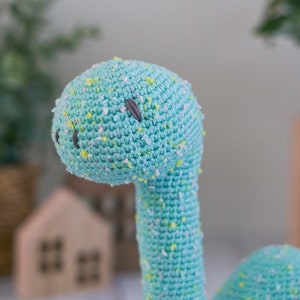 Puede incluir: Un juguete de dinosaurio azul de crochet con textura moteada y ojos negros. El dinosaurio est&aacute; de pie con la cabeza inclinada hacia un lado.
