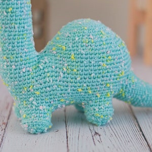 Puede incluir: Un juguete de dinosaurio de crochet azul claro con motas blancas y amarillas. El dinosaurio est&aacute; de pie sobre una superficie de madera blanca.