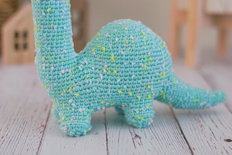 Puede incluir: Un juguete de dinosaurio de crochet azul claro con motas blancas y amarillas. El dinosaurio est&aacute; de pie sobre una superficie de madera blanca.