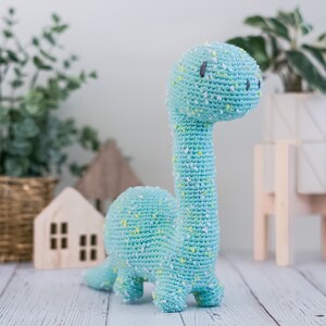 Puede incluir: Un juguete de dinosaurio de crochet azul claro con motas amarillas. El dinosaurio tiene un cuello largo y una cabeza peque&ntilde;a con ojos negros.