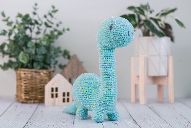 Puede incluir: Un juguete de dinosaurio de crochet azul claro con motas amarillas. El dinosaurio tiene un cuello largo y una cabeza peque&ntilde;a con ojos negros.