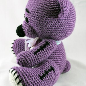 May include: A purple crocheted teddy bear with black eyes and stitched details. The bear has white stitching around the edges of its paws and ears.