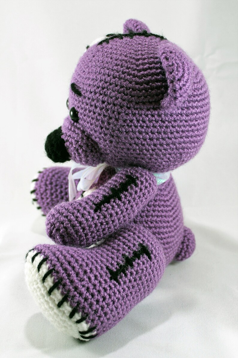 May include: A purple crocheted teddy bear with black eyes and stitched details. The bear has white stitching around the edges of its paws and ears.