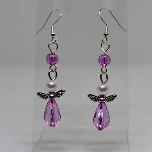 May include: A pair of dangle earrings featuring a silver angel charm, a white pearl bead, and a pink faceted teardrop bead. The earrings are suspended from silver hooks.