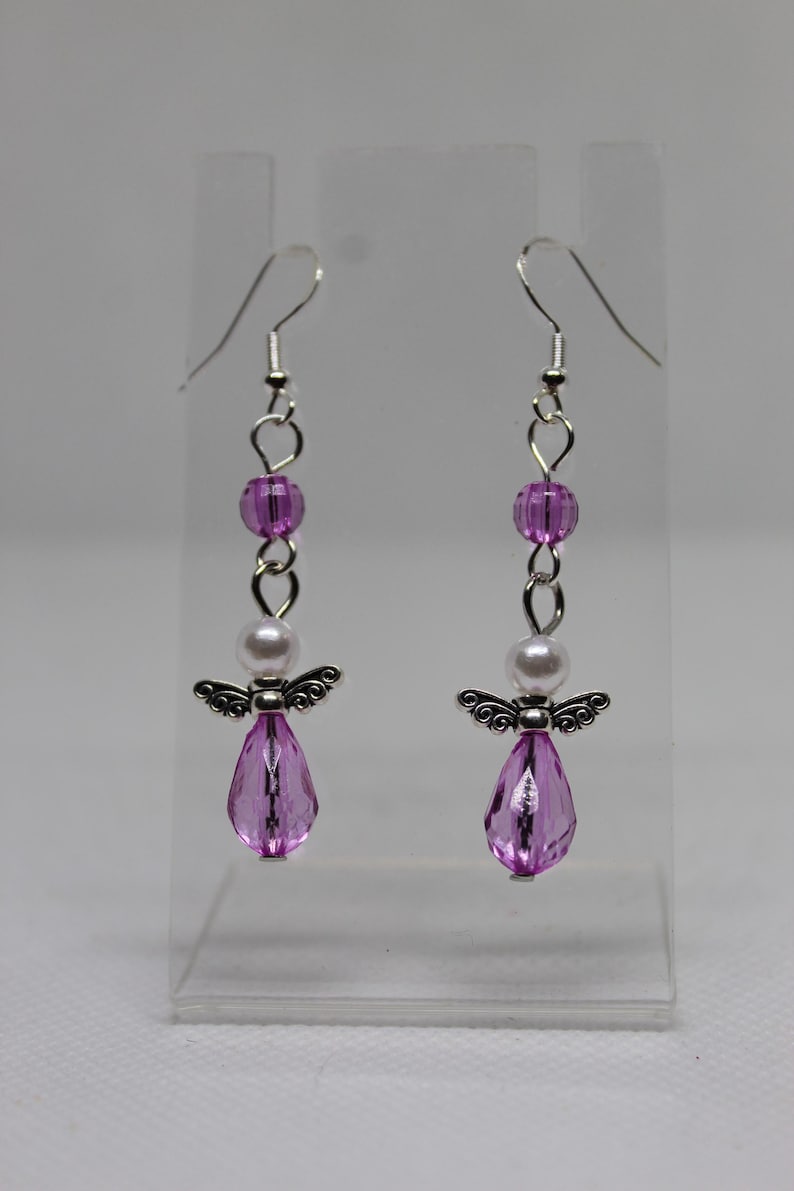May include: A pair of dangle earrings featuring a silver angel charm, a white pearl bead, and a pink faceted teardrop bead. The earrings are suspended from silver hooks.