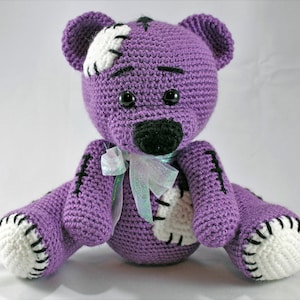 May include: A purple crocheted teddy bear with white patches and black stitching. The bear has black eyes and a black nose. It is wearing a light blue and white ribbon around its neck.