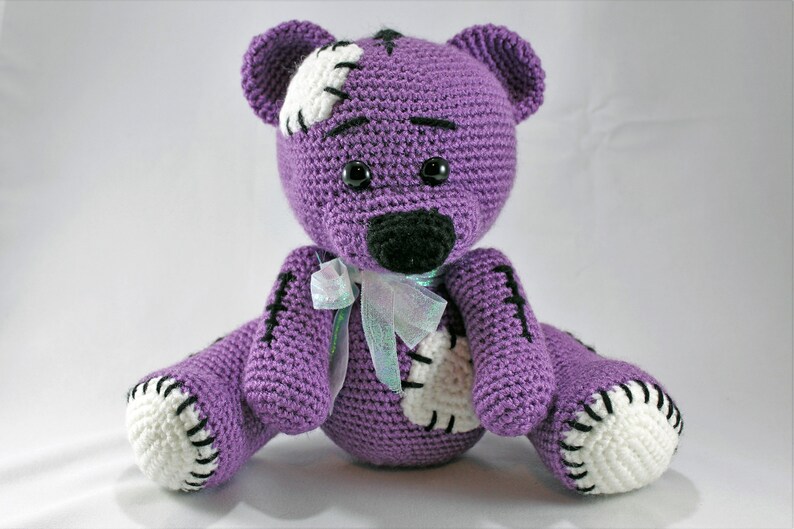 May include: A purple crocheted teddy bear with white patches and black stitching. The bear has black eyes and a black nose. It is wearing a light blue and white ribbon around its neck.