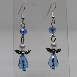 May include: A pair of dangle earrings featuring a silver angel charm, a white pearl bead, and a light blue crystal teardrop. The earrings are suspended from silver hooks.