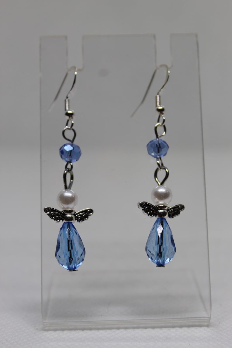 May include: A pair of dangle earrings featuring a silver angel charm, a white pearl bead, and a light blue crystal teardrop. The earrings are suspended from silver hooks.