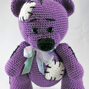 May include: A purple crocheted teddy bear with white and black accents. The bear has a stitched patch on its chest and a white bow around its neck.