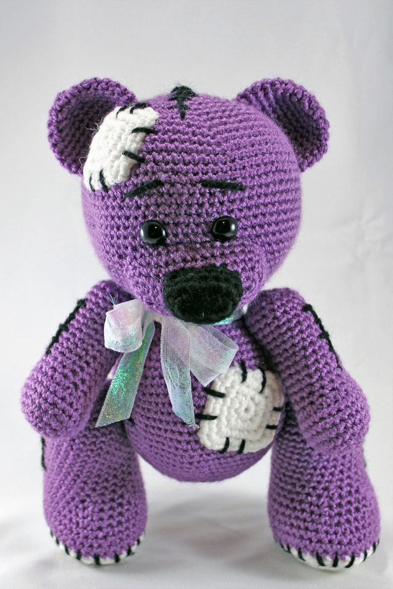 May include: A purple crocheted teddy bear with white and black accents. The bear has a stitched patch on its chest and a white bow around its neck.