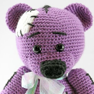 May include: Purple crocheted teddy bear with black eyes and a black nose. The bear has a white patch on its head with black stitching.