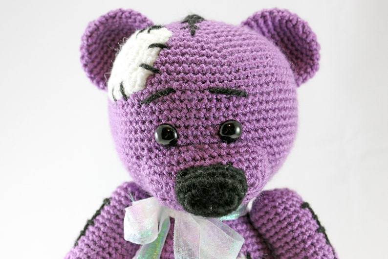May include: Purple crocheted teddy bear with black eyes and a black nose. The bear has a white patch on its head with black stitching.