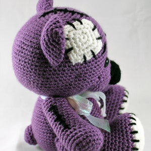 May include: Purple crocheted teddy bear with white and black stitching details. The bear has a white patch on its head and a white ribbon around its neck.