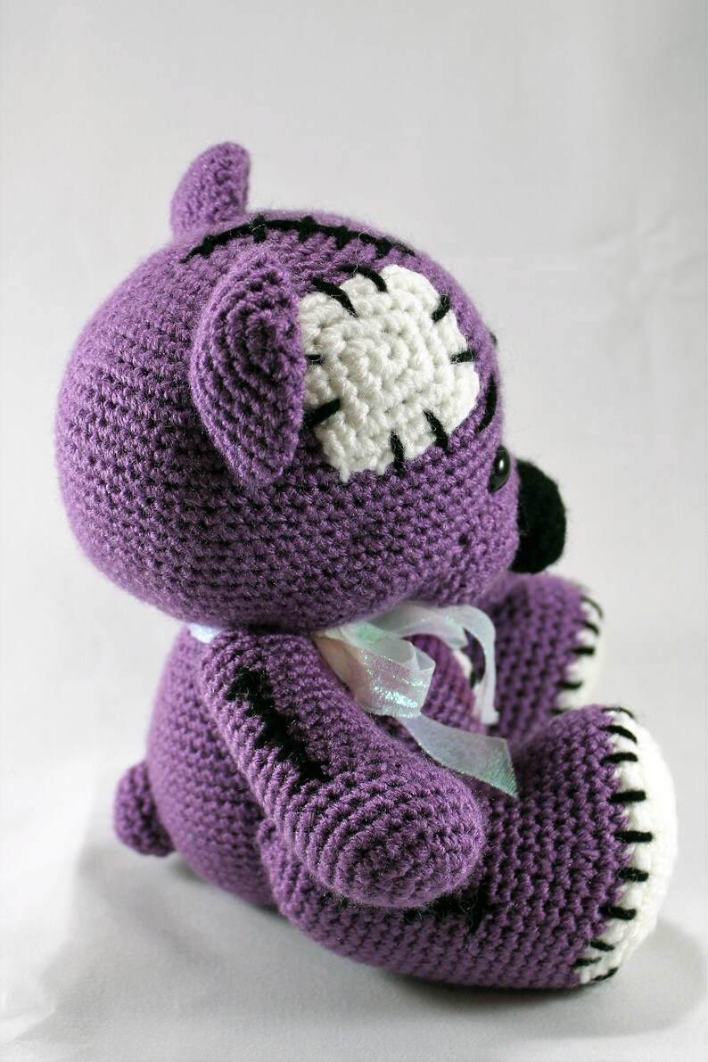 May include: Purple crocheted teddy bear with white and black stitching details. The bear has a white patch on its head and a white ribbon around its neck.