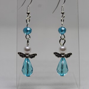 May include: A pair of dangle earrings featuring a silver angel charm with a light blue crystal teardrop, a white pearl bead, and a light blue crystal bead.
