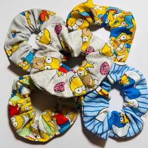 May include: Five fabric scrunchies featuring Homer Simpson from The Simpsons cartoon. The scrunchies are in various colors, including gray, yellow, and blue, with donut and character prints. These hair accessories are perfect for adding a touch of fun to any hairstyle.