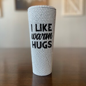 Op de afbeelding: Een witte beker bedekt met kleine, ronde, parelachtige versieringen. De tekst "I LIKE warm HUGS" is in zwarte, gestileerde letters op de voorkant van de beker geschreven. De beker is cilindrisch.