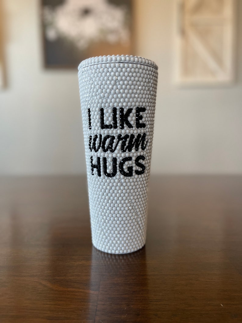 Op de afbeelding: Een witte beker bedekt met kleine, ronde, parelachtige versieringen. De tekst "I LIKE warm HUGS" is in zwarte, gestileerde letters op de voorkant van de beker geschreven. De beker is cilindrisch.