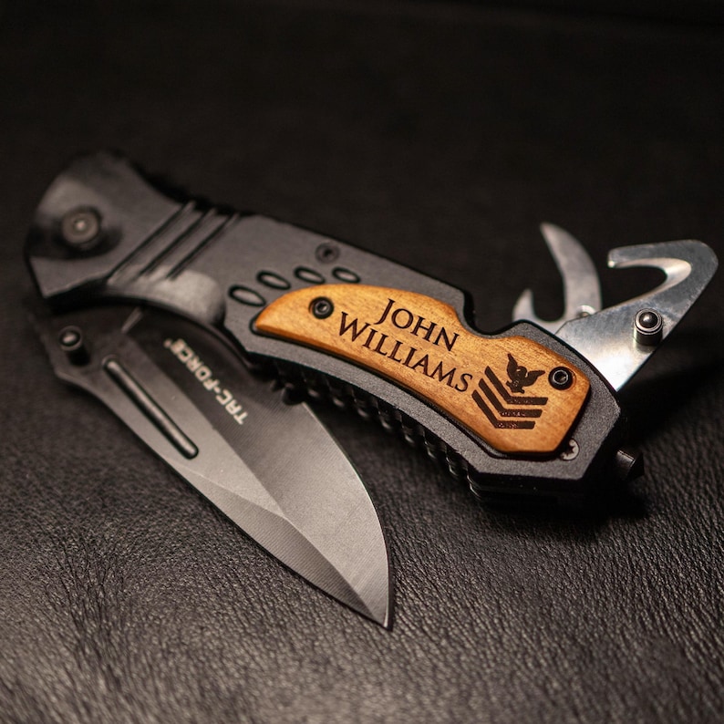May include: A black multi-tool pocket knife with a wooden handle. The handle is engraved with the name "JOHN WILLIAMS" and a military insignia. The knife has a blade, a bottle opener, a saw, and a screwdriver.