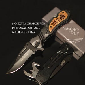 May include: A black folding knife with a wood handle engraved with the name "Daniel Stone". The knife is open and the blade is visible. The knife is in a box with the words "Smoky Tree" printed on it.