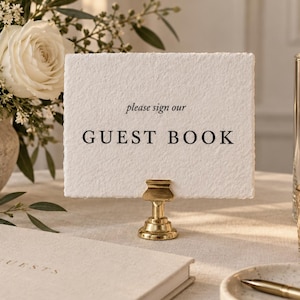 Puede incluir: Una tarjeta rectangular blanca con bordes irregulares sostenida por un soporte de metal dorado, con el texto impreso en negro "please sign our GUEST BOOK". La tarjeta está junto a un libro de visitas beige, un bolígrafo dorado y una vela.