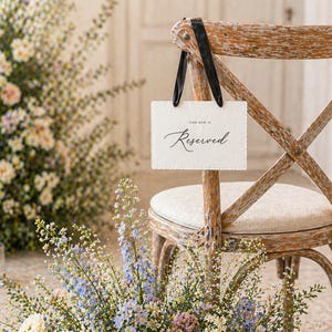 May include: A rustic wooden chair with a sign that reads "Reserved" hangs from the back. The chair is surrounded by a floral arrangement of white, pink, and blue flowers. Two candles in glass holders are in the background.