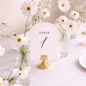 May include: A table setting with white cosmos flowers and baby's breath in small glass vases. A white card with the word "TABLE" and the number "1" is held by a gold clip. A candle in a glass holder is also on the table.