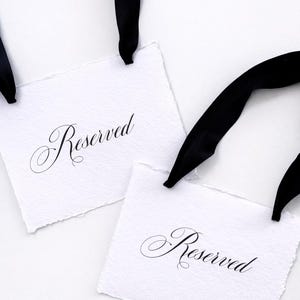 Op de afbeelding: Twee witte papieren labels met het woord "Reserved" in elegant schrift, elk bevestigd aan een zwart satijnen lint. De labels hebben een getextureerde, gescheurde rand en de linten zijn bovenaan gelust. De achtergrond is effen wit.
