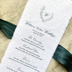 May include: A wedding invitation with a rustic, hand-torn paper texture. The invitation features a simple wreath design with the initials "K" and "M" in the center. The text reads "Welcome to our Wedding, Kaitlin & Marcus, April 18, 2022". The invitation lists the wedding ceremony, cocktail hour, dinner, and dancing times.