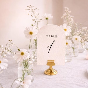 May include: A table number card with the word "TABLE" and the number "1" in black script. The card is held by a gold-colored metal stand. White flowers and candles are in the background, creating a soft, romantic ambiance.