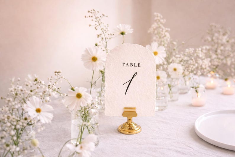 May include: A table number card with the word "TABLE" and the number "1" in black script. The card is held by a gold-colored metal stand. White flowers and candles are in the background, creating a soft, romantic ambiance.