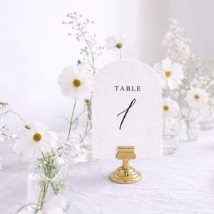 Puede incluir: Decoración de mesa de boda con una tarjeta blanca que dice "TABLE 1" en escritura negra, sujeta por un clip dorado. Flores blancas y pequeños jarrones de cristal con flores están dispuestas alrededor de la tarjeta.