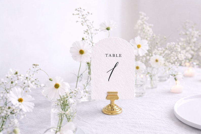 May include: A wedding table setting with a white card that reads "TABLE 1" in black script, held by a gold-colored clip. White flowers and small glass vases with flowers are arranged around the card. The table is covered with a white tablecloth.