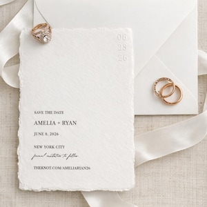 以下が含まれることがあります： SAVE THE DATE、AMELIA + RYAN、JUNE 8, 2026、NEW YORK CITY、formal invitation to follow、THEKNOT.COM/AMELIARYAN26と黒い文字が印刷された白い手漉き風のセーブ・ザ・デートカード。白い封筒、リボン、ゴールドの指輪が添えられています。