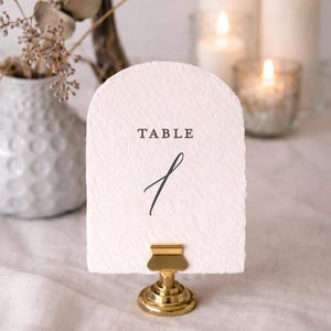 May include: A white, arched table number card with the word "TABLE" and the number "1" in black script. The card is held by a gold-colored metal clip. Candles and a vase with dried flowers are in the background.