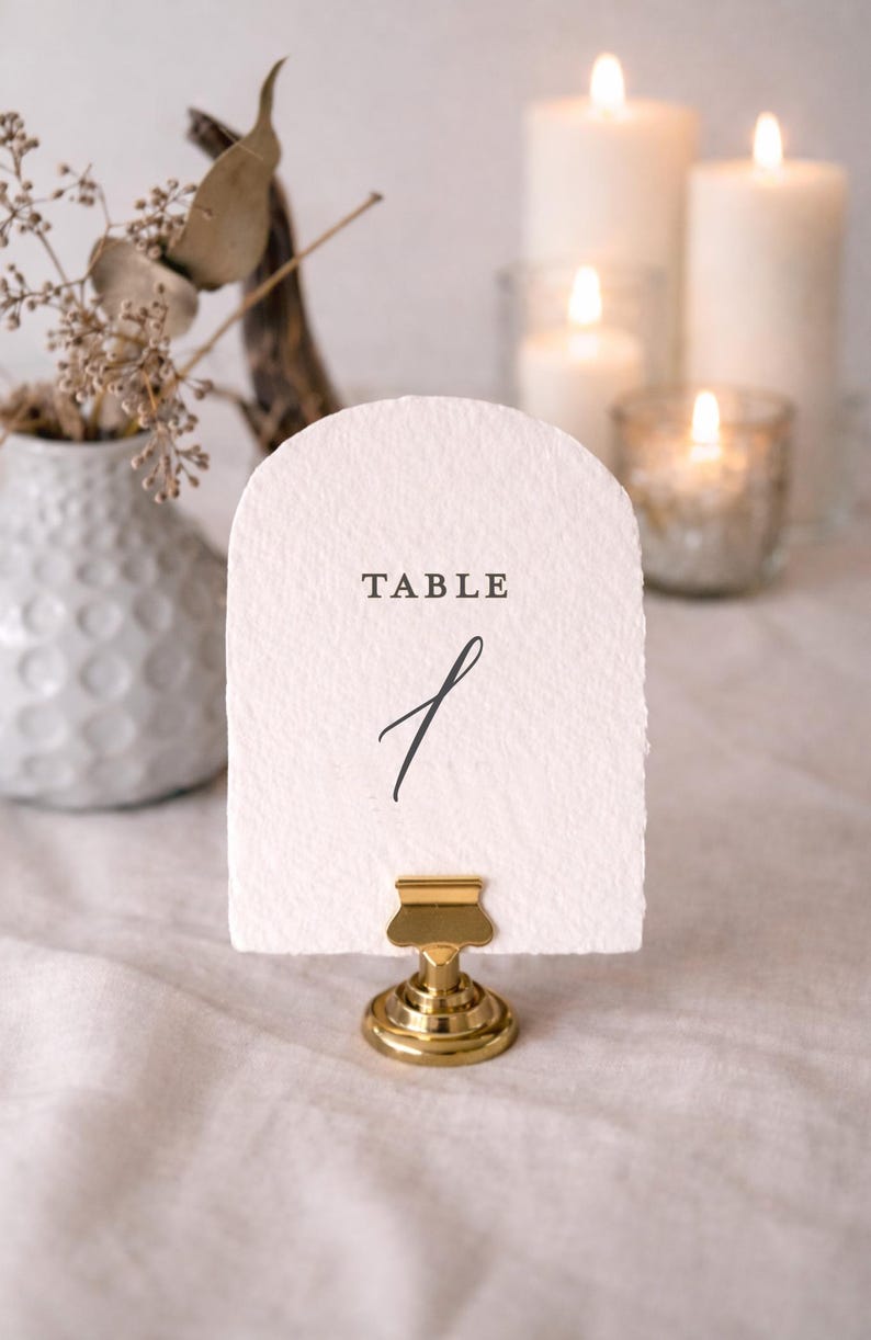 May include: A white, arched table number card with the word "TABLE" and the number "1" in black script. The card is held by a gold-colored metal clip. Candles and a vase with dried flowers are in the background.