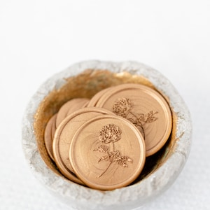 May include: A small bowl filled with gold wax seals featuring a floral design. The seals are round and have a raised design of a flower with leaves.