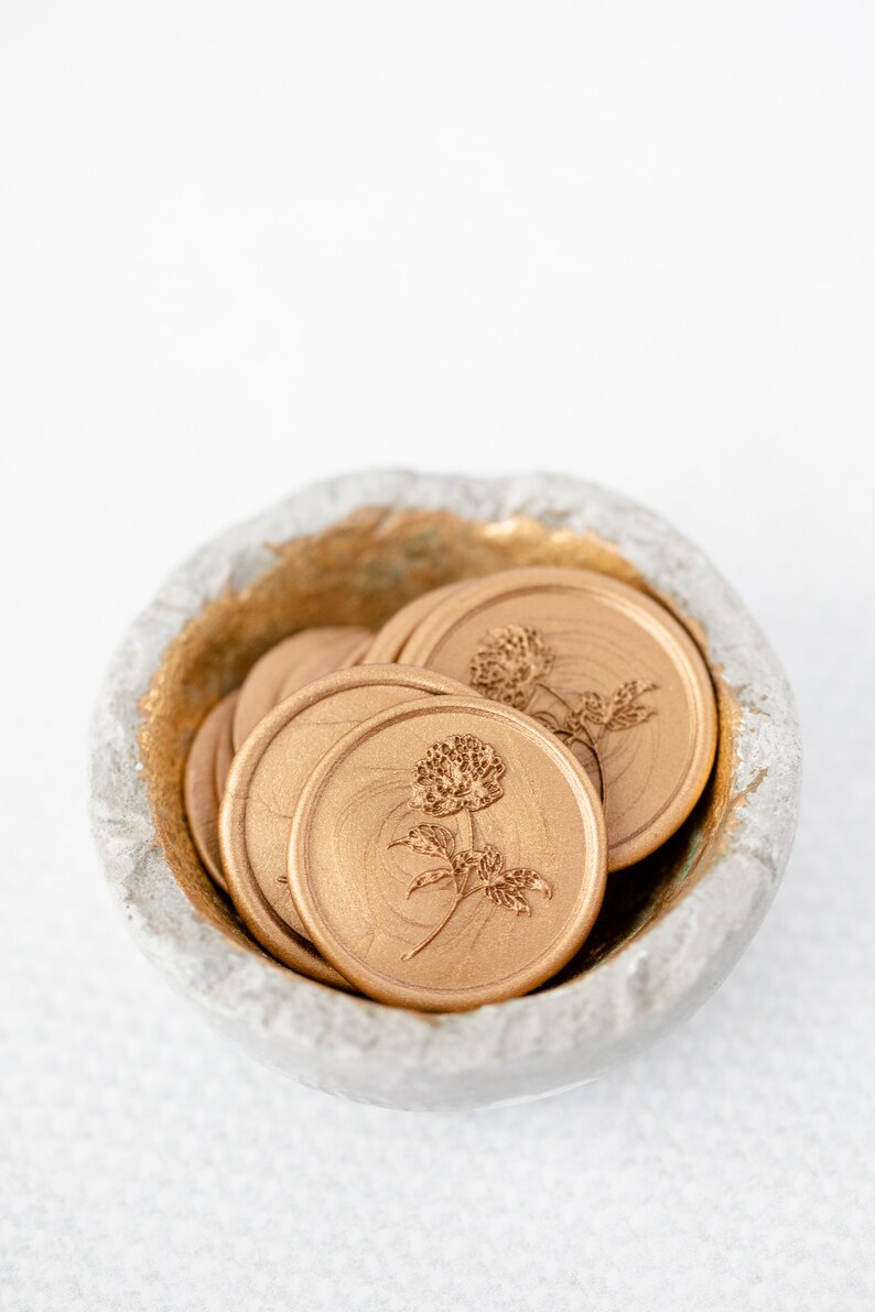 May include: A small bowl filled with gold wax seals featuring a floral design. The seals are round and have a raised design of a flower with leaves.