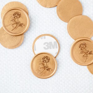 May include: Gold wax seals with a floral design. The seals are on a white surface. Some of the seals have a 3M adhesive backing.