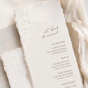 May include: Two textured, off-white wedding menu cards with torn edges. The cards display the date "06 04 23" and the phrase "eat, drink, be married." The menu lists a Caesar salad, chicken piccata, and filet mignon.
