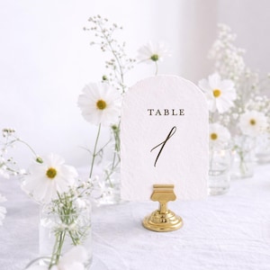 May include: An arched, textured white paper table number card with the word TABLE and the number 1 printed in black. The card is held upright by a small, polished gold-colored metal stand with a circular base, positioned on a white surface.