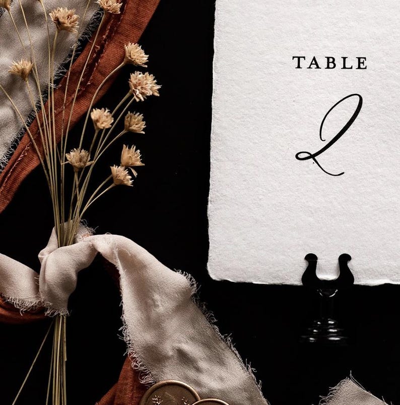 May include: A still-life image featuring a bouquet of dried flowers, a white paper with the word "TABLE" and the number "2", and a black metal table number holder. The scene includes a beige ribbon and a dark background.