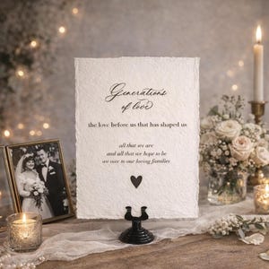 May include: A wedding reception table setting with a framed black and white photo, a sign with the text "Generations of love", candles, and a floral arrangement. The sign is held by a black metal stand.