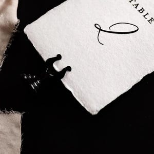 May include: A black metal place card holder gripping a white paper card with the word "TABLE" and a decorative script initial. A piece of beige fabric is in the background. The background is black.